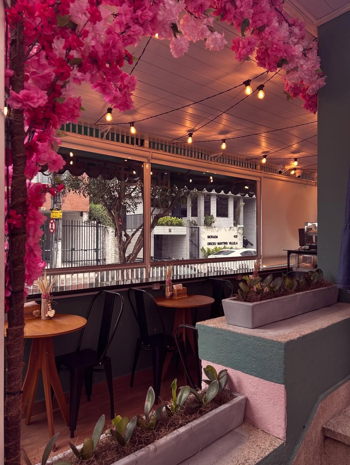 Varanda do Le Petit Café com galhos de flores cor-de-rosa, mesas de madeira, cadeiras pretas e varal de lâmpadas acesas.