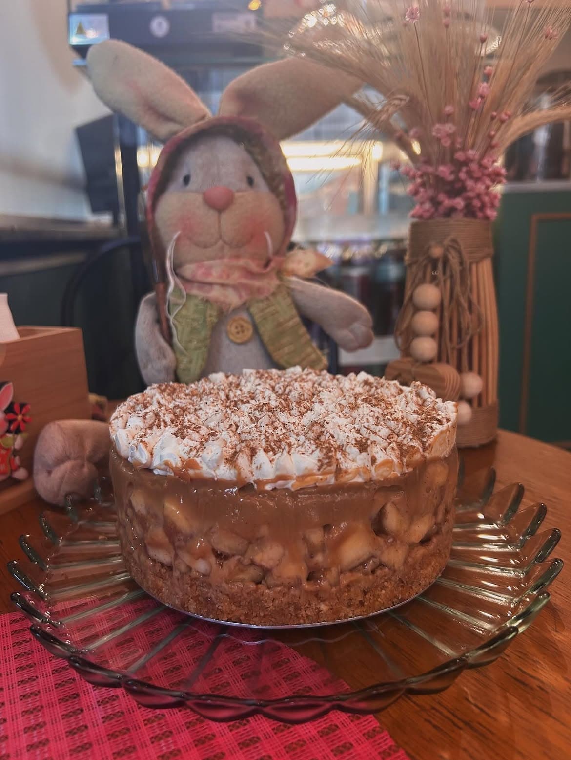 Coelho de pano com lenço rosado e vaso de palha com flores secas sobre uma mesa.