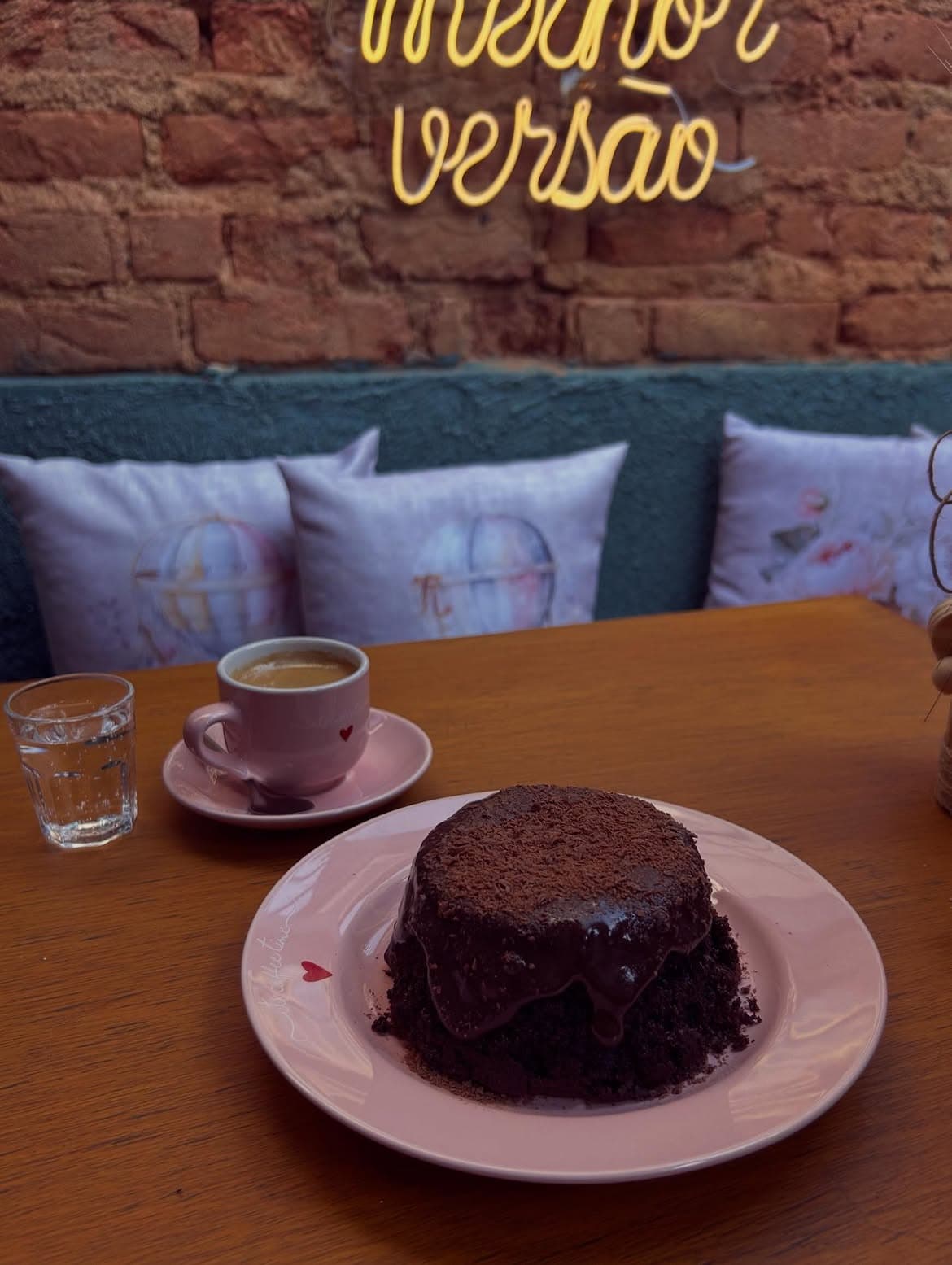 Bolo de chocolate com calda escorrendo, ao lado de uma xícara rosa de café, diante de um neon com a frase “melhor versão”.
