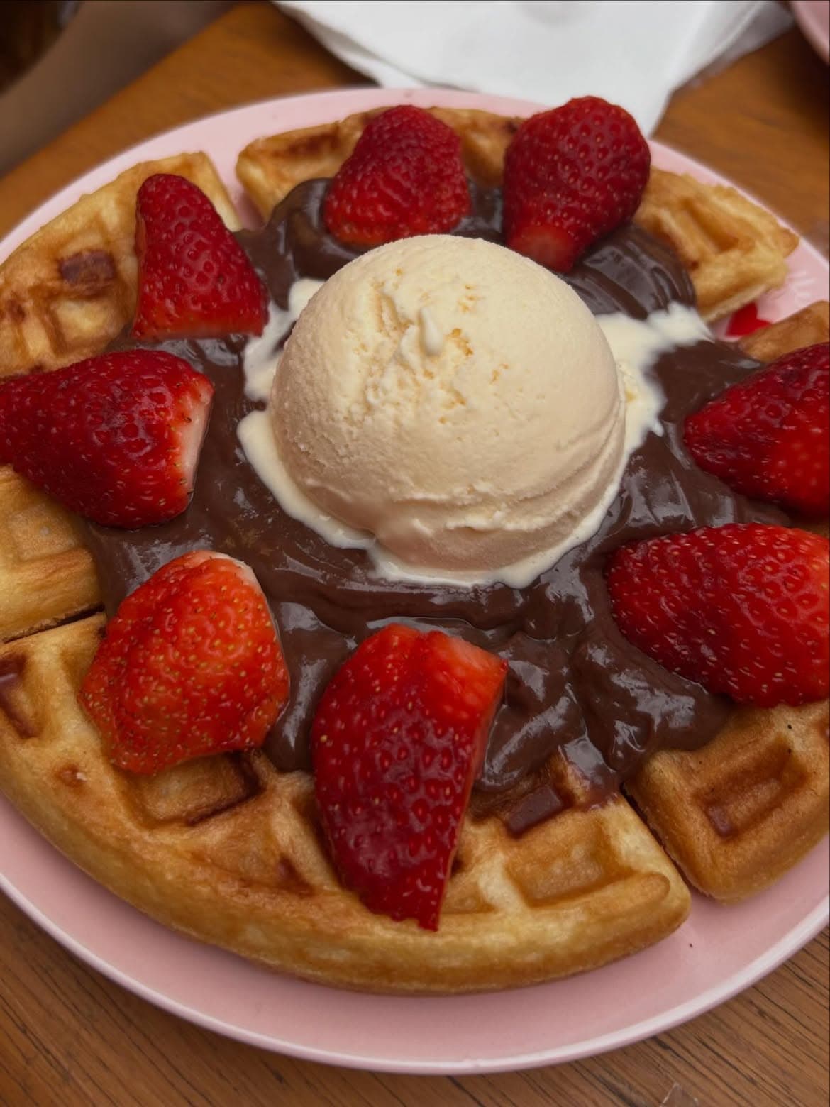 Waffle com creme de chocolate, morangos frescos e uma bola de sorvete de creme em prato rosa.